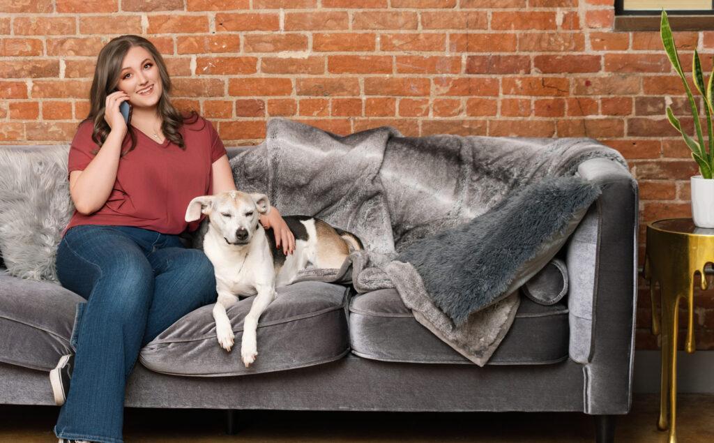 Allie in a red shirt and jeans, on a gray couch. She is talking on the phone, smiling at us, and petting a black, brown, and white dog.