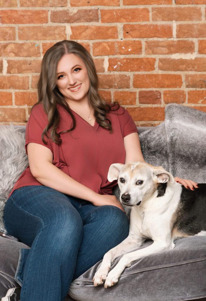 Allie Wickam in a red blouse and blue jeans on a gray couch with a brown and black dog in front of a brick wall